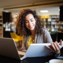 Eine junge Frau sitzt in einem Buchladen am Laptop und schaut währenddessen in ein Buch. 