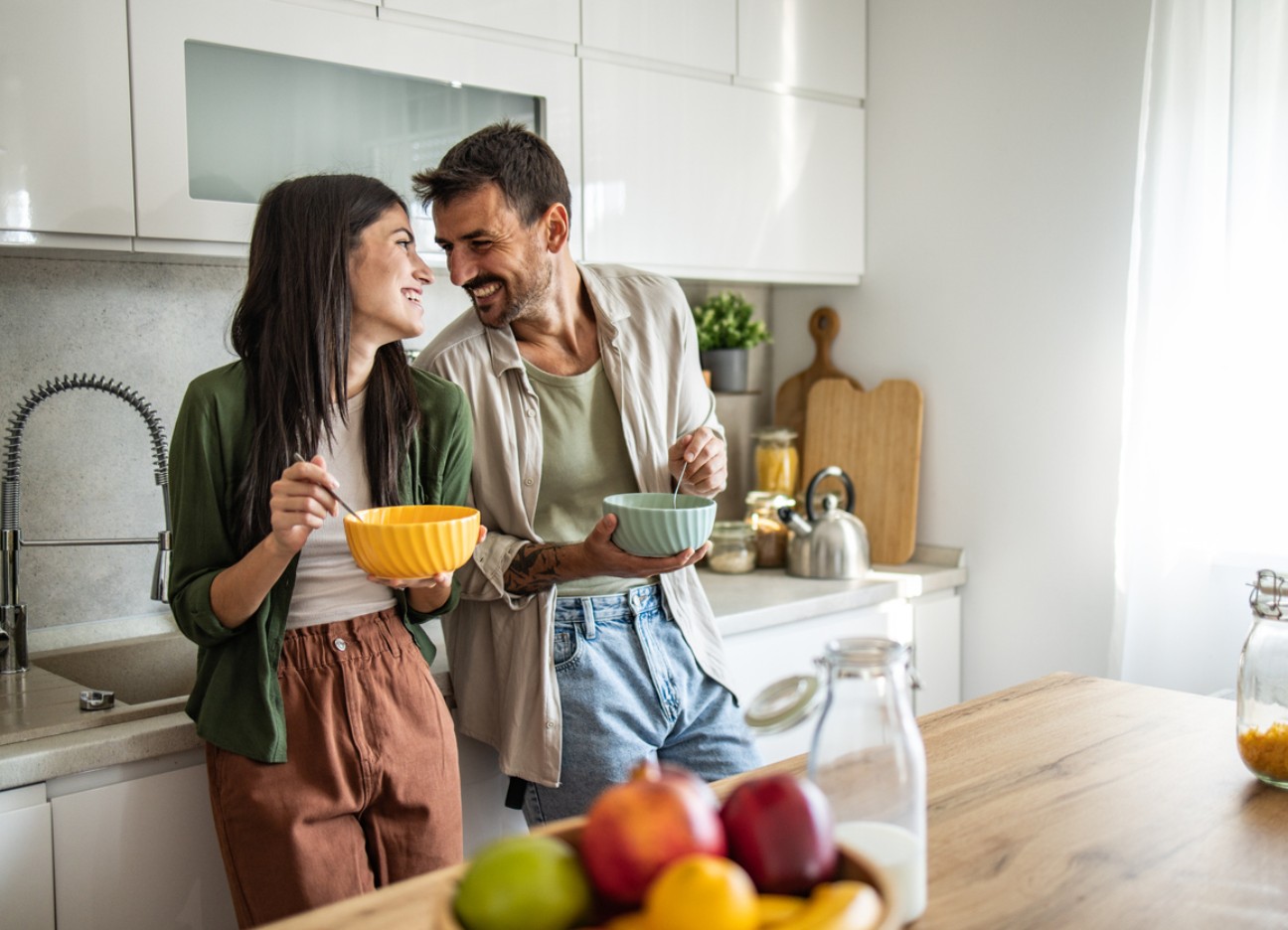 Ein junges Paar steht mit Müslischalen in der Hand steht lachend in der Küche