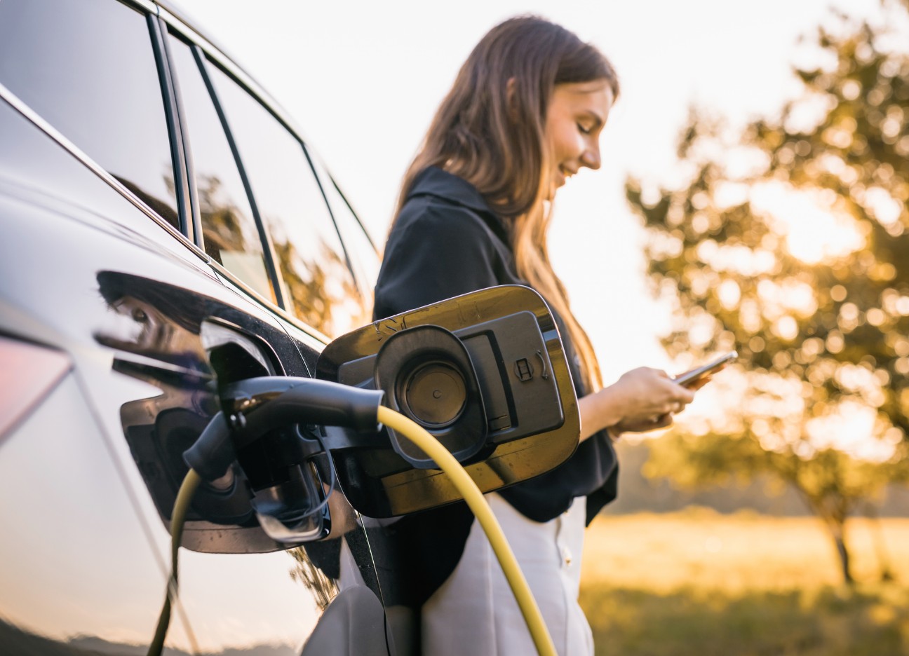 Bühnenbild Ratgeber Elektroauto Eine Frau steht neben einem Elektroauto, das gerade aufgeladen wird und schaut auf ihr Smartphone.
