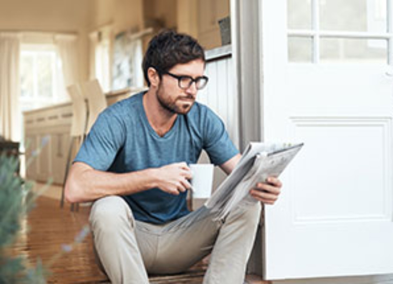 Person sitzt auf der Türschwelle, hält eine Zeitung und eine Tasse.