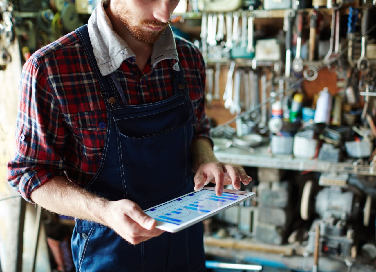 Ein Handwerker steht in einer Garage mit Handwerksequipment und schaut auf ein Tablet. 