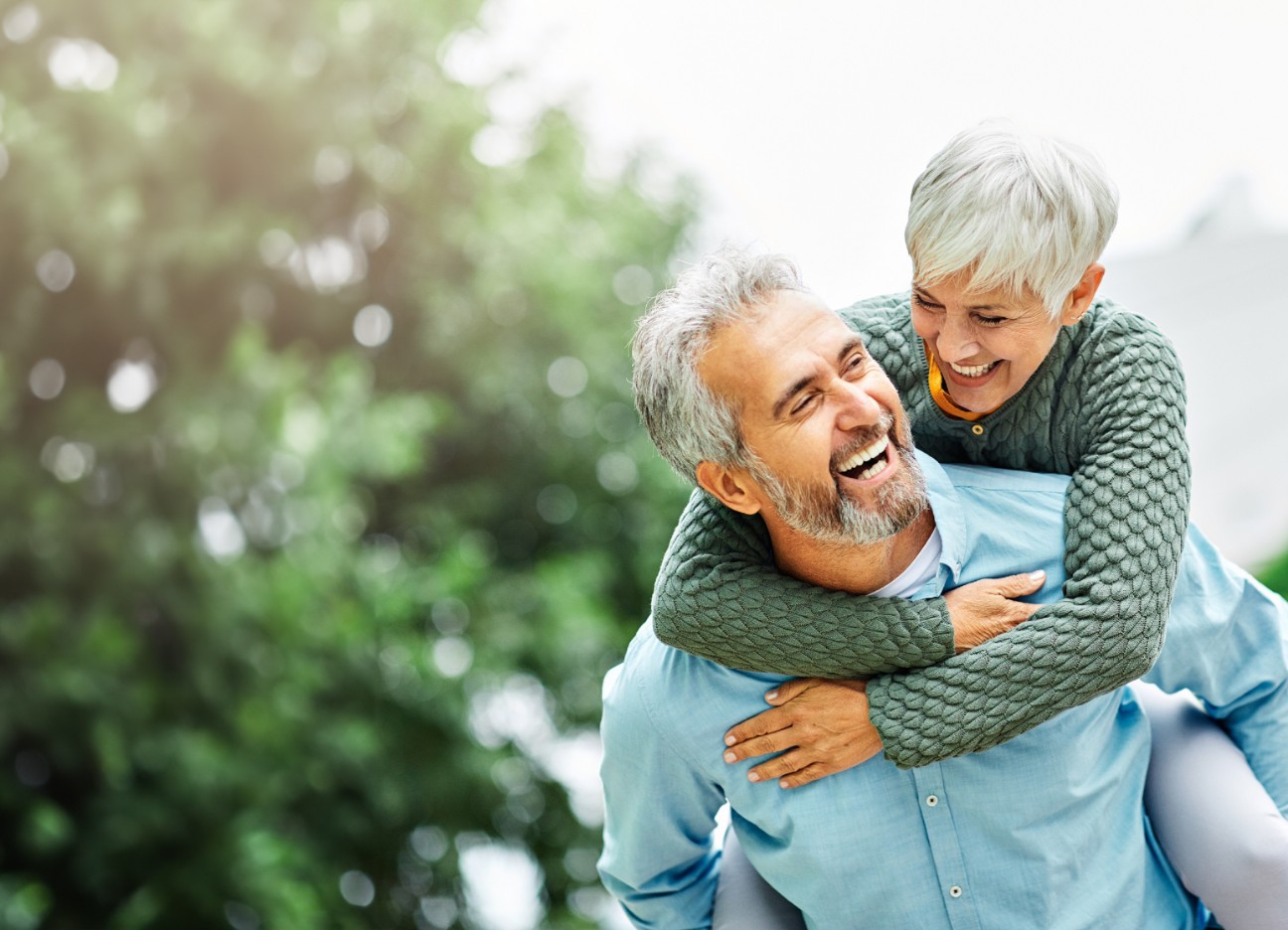 Ein älteres Paar läuft lachend durch den Park. Der Mann trägt die Frau huckepack.