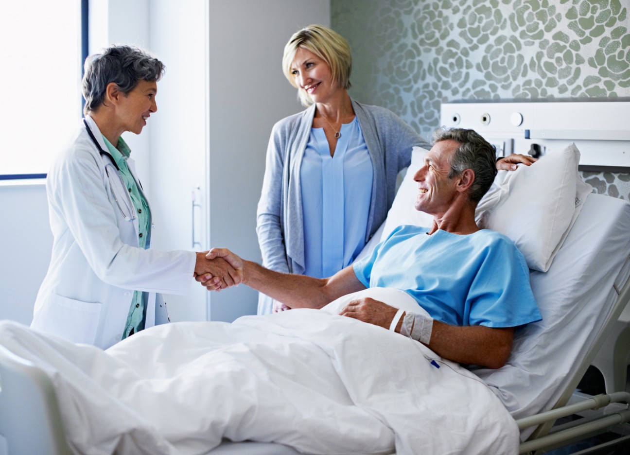 Ein Patient im Krankenhausbett schüttelt eine Ärztin die Hand, während seine Frau daneben steht.