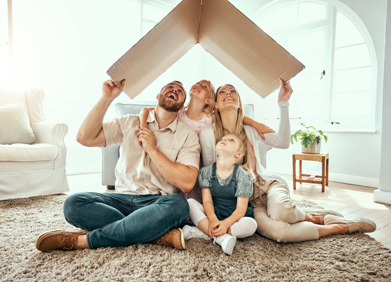 Familie sitzt lachend unter einem Dach aus Karton.