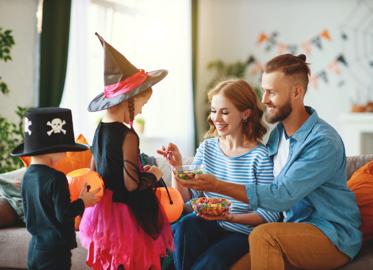 Bühnenbild Ratgeber Halloween Zwei auf dem Sofa sitzende Eltern geben ihren verkleideten Kindern Süßigkeiten in ihren Halloween-Eimer.