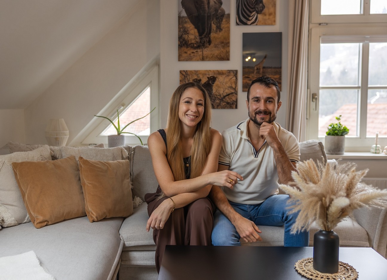 Junges Paar sitzt nebeneinander auf einem weißen Ecksofa in einer Dachgeschoßwohnung und lacht in die Kamera