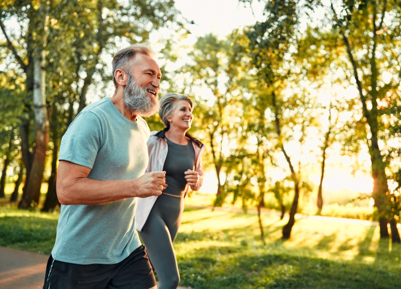 Bühnenbild Ratgeber gesundes Herz Vorsorge Ein älteres Paar joggt gemeinsam lachend durch einen sonnigen Park.
