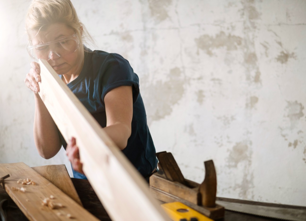 Bühnenbild Ratgeber Gefahren auf der Baustelle Eine Frau arbeitet an einem länglichen Stück Holz. Sie trägt eine Schutzbrille und neben ihr liegen diverse Werkzeuge.
