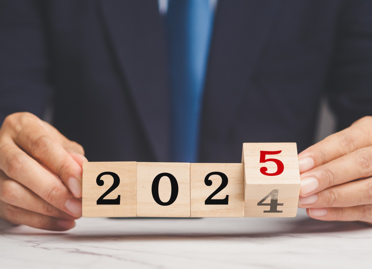 Bühnenbild Ratgeber Neue Gesetze 2025 Eine Person mit Anzug hält Holzklötze mit den Zahlen 2024 und 2025 in der Hand. Die Zahl 4 wird hierbei durch eine 5 ersetzt.