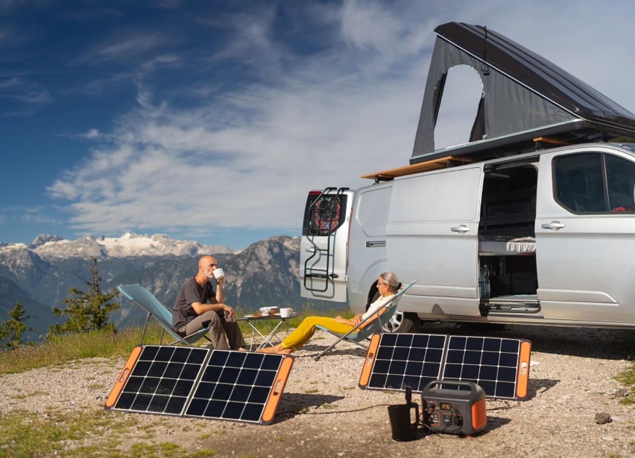 Bühnenbild Ratgeber Strom zum Heizen selbst erzeugen: Geht das? Eine Frau und ein Mann sitzen zusammen in einem Liegestuhl vor ihrem Campervan in den Bergen. Vor ihnen stehen zwei Solarpanels.
