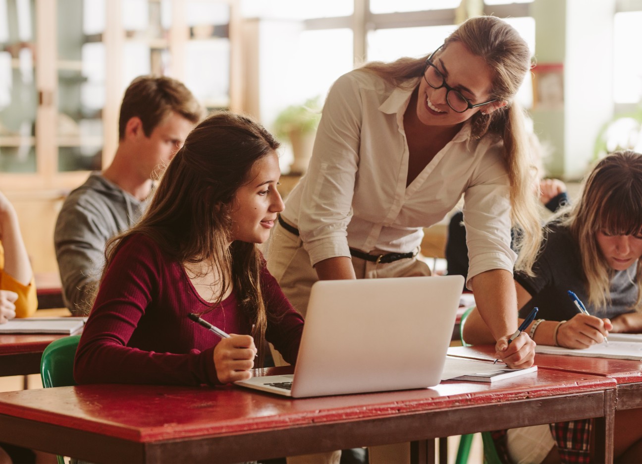 Eine Lehrerin hilft einer Schülerin, die an ihrem Tisch im Klassenraum arbeitet.