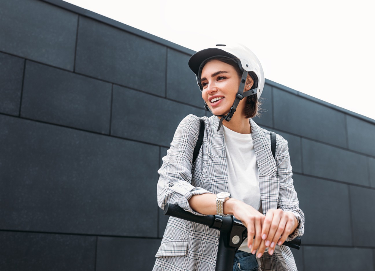 Person mit Helm und kariertem Blazer steht neben einem E-Scooter vor einer dunklen Wand.