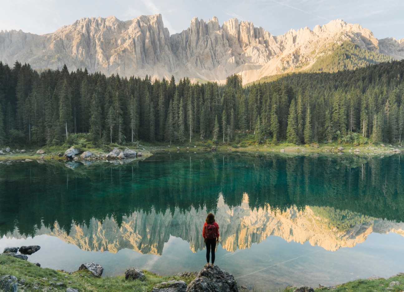 Person in roter Jacke steht am Ufer eines Sees, der von Bergen und Wäldern umgeben ist. Person in roter Jacke steht am Ufer eines Sees, der von Bergen und Wäldern umgeben ist.