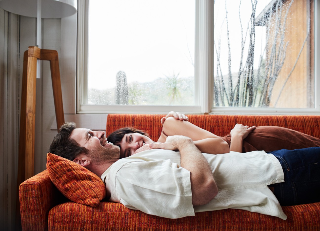 Mann und Frau liegen aneinander gekuschelt auf einem Sofa während es draußen stark regnet Mann und Frau liegen aneinander gekuschelt auf einem Sofa während es draußen stark regnet
