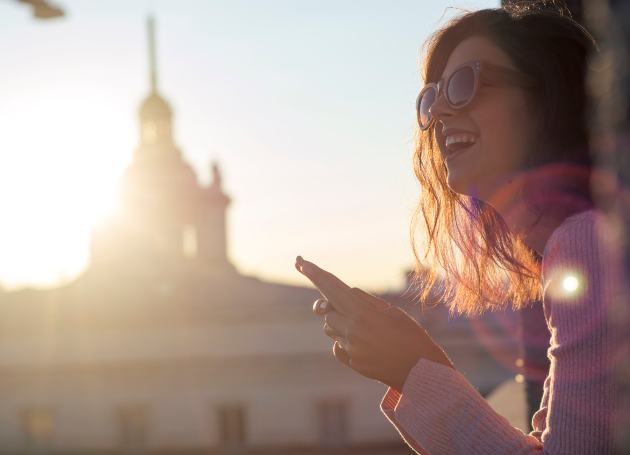 Bühnenbild Ratgeber Reiseversicherung Studenten Eine junge Frau schaut lächelnd von ihrem Smartphone hoch, während sie vor einem größeren, weißen Gebäude sitzt.
