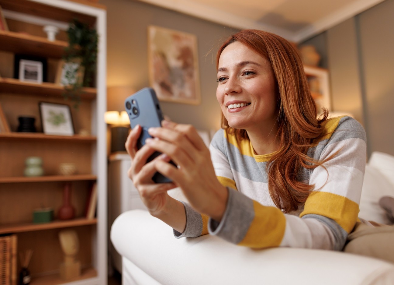 Eine junge Frau sitzt auf einem Sofa und schaut lachend auf ihr Smartphone.