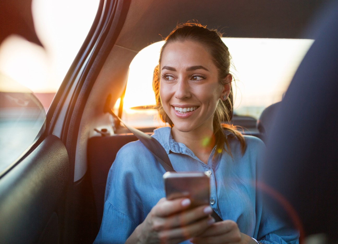 Bühnenbild Ratgeber Digitale Revolution Auswirkungen auf Leben und Arbeitswelt Frau sitzt auf der Rückbank, schaut lachend aus dem Fenster hinaus während sie ein Telefon in der Hand hält.