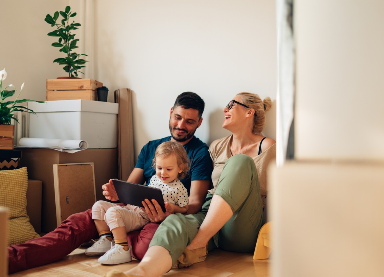 Eine Familie sitzt zwischen Umzugskartons auf dem Boden; ein Mann und ein kleines Kind schauen gemeinsam auf ein Tablet, während eine Frau daneben lachend zusieht.