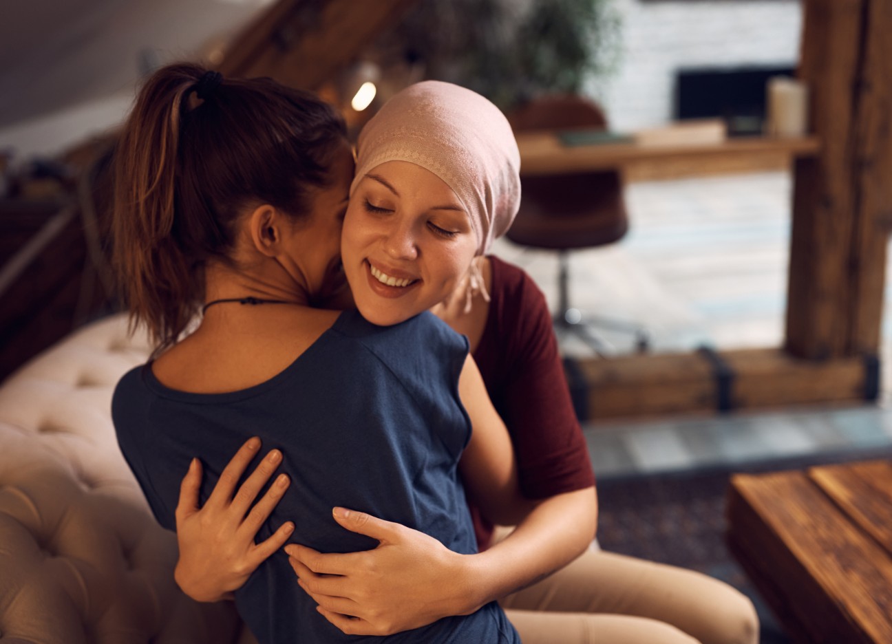Bühnenbild Ratgeber Krebsversicherung Zwei junge Frauen sitzen auf dem Sofa und umarmen sich. Eine der Frauen trägt hat ein Tuch auf dem Kopf.