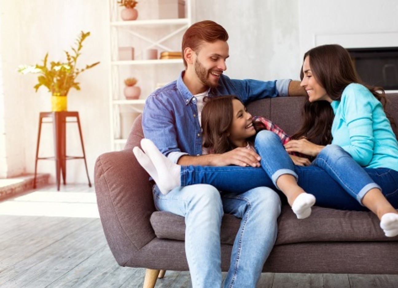 Bühnenbild Ratgeber Erfindungen für einen sicheren Haushalt Zwei junge Eltern sitzen lachend mit ihrer Tochter auf dem Sofa.