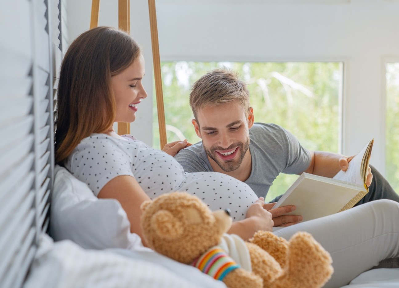 Bühnenbild Ratgeber Absicherung für Babys Eine hochschwangere Frau liegt aufrecht im Bett, neben ihr ein Teddybär und ein Mann liest ein Buch vor, während er auf den Babybauch schaut.