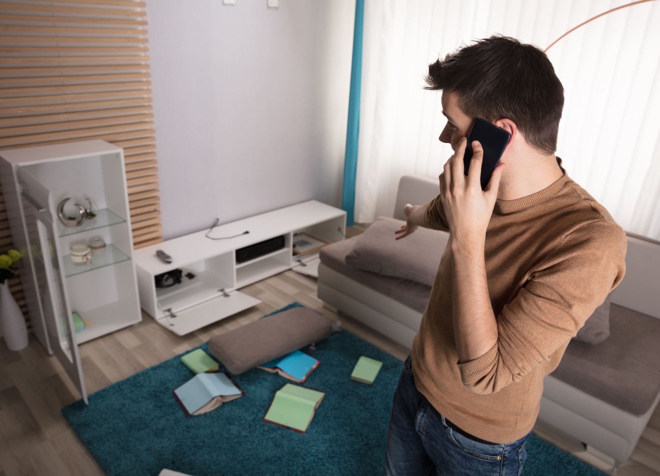 Bühnenbild Ratgeber Einbruch was tun? Ein junger Mann telefoniert, während er in seinem verwüsteten Wohnzimmer steht.