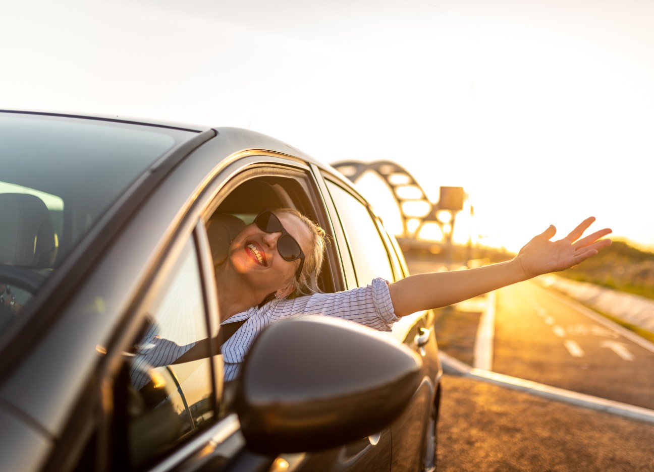 Eine junge blonde Frau fährt in glücklich in der Sonne ihr Auto, trägt eine Sonnenbrille, streckt einen Arm aus dem Fahrerfenster und lacht.