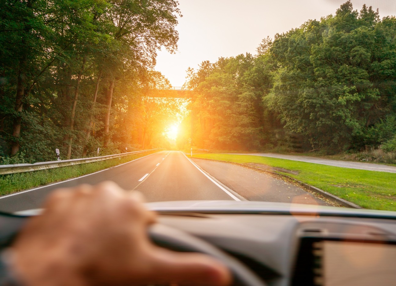 Bühnenbild Ratgeber CBD und Autofahren Eine zusehende Hand am Lenkrad eines Autos. Das Auto fährt eine Landstraße im Sonnenuntergang entlang.