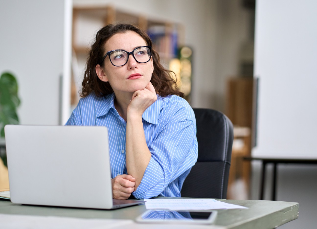 Bühnenbild Ratgeber Organspendeausweis Nachdenkliche Frau mit Brille sitzt am Schreibtisch vor einem Laptop, stützt ihr Kinn auf die Hand und schaut zur Seite.