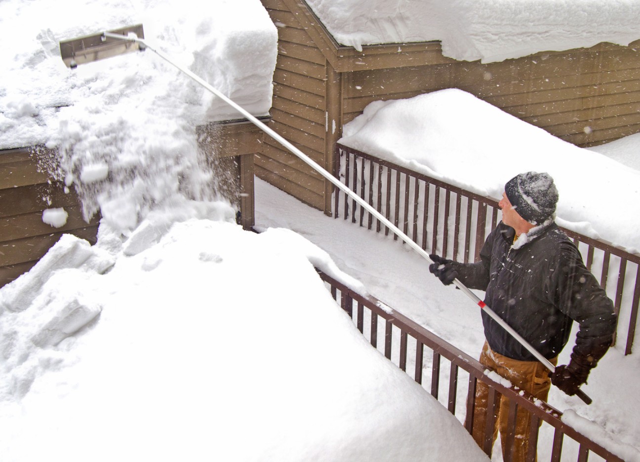 Bühnenbild Ratgeber Elementarschäden Ein Mann mit einem Schnee-Rechen entfernt Schnee vom Dach.