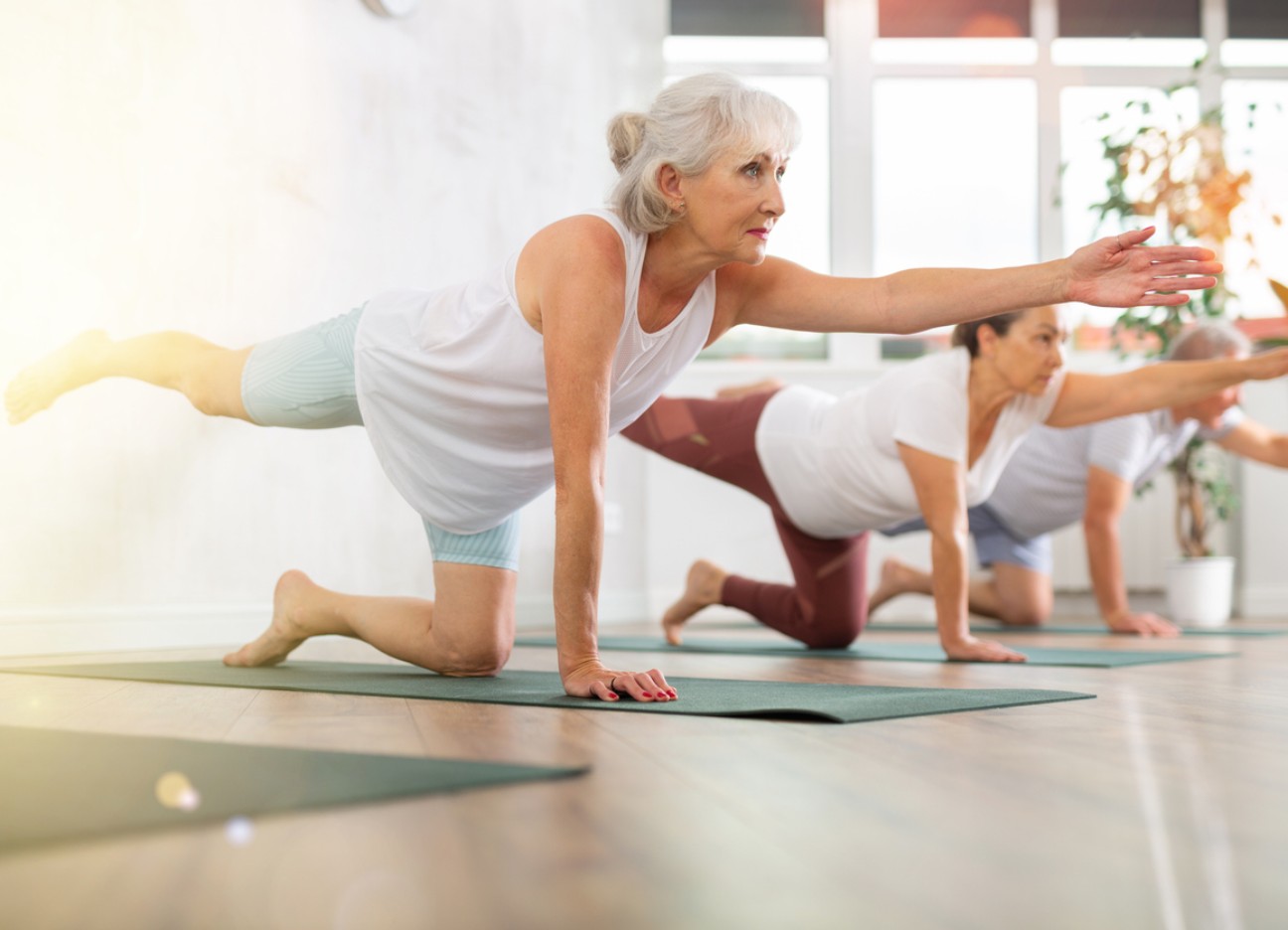 Bühnenbild: Ratgeber stuerze im alter sturzpraevention Ältere Frau macht mit anderen eine Balance-Übung auf Yogamatten in einem hellen Raum.