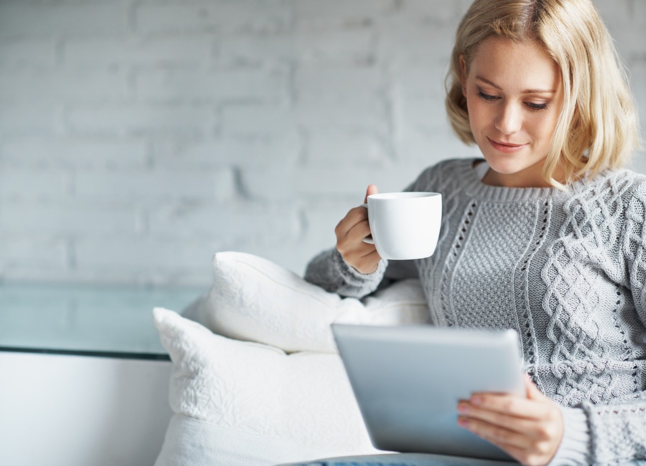 Person sitzt auf einem Sofa, hält eine Tasse und ein Tablet, trägt einen grauen Pullover.