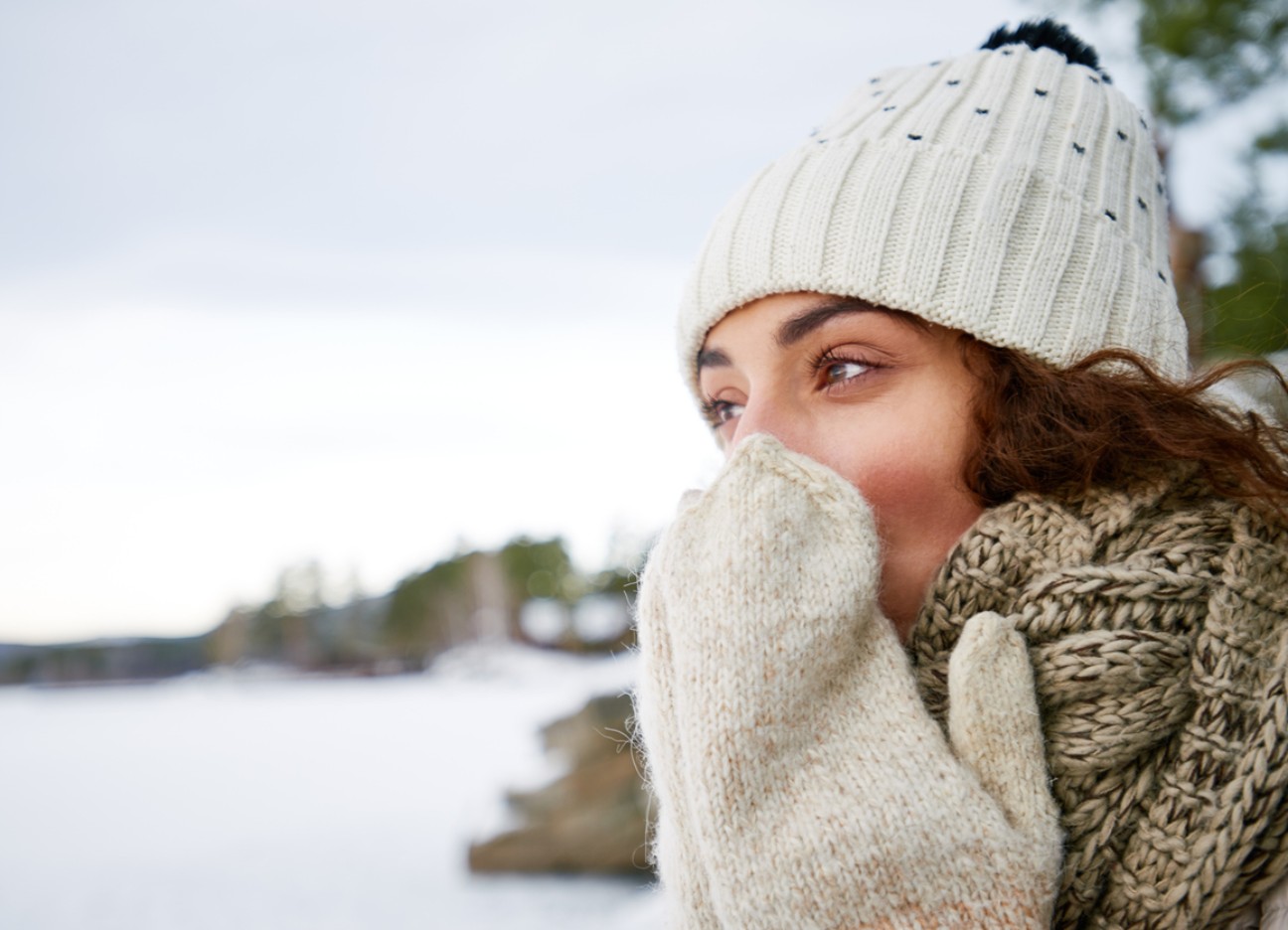 Eine warm eingepackte Frau mit Mütze, Schal und Handschuhen steht draußen in einer verschneiten Landschaft und hält die Hände vor ihr Gesicht, um sich zu wärmen.