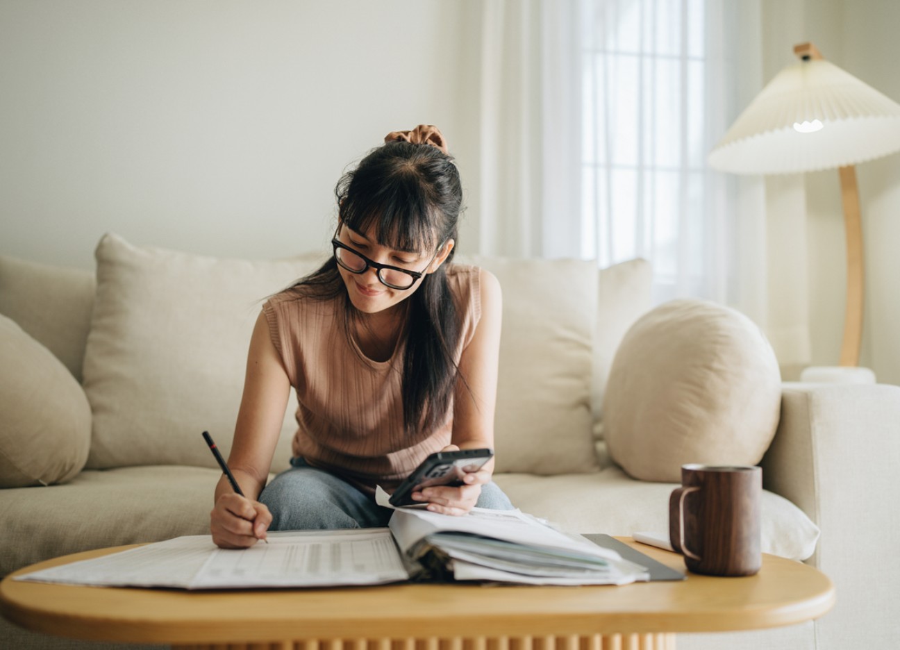 Eine junge Frau sitzt auf einem Sofa, schaut auf Unterlagen auf dem Tisch und macht Notizen, während sie ein Smartphone in der Hand hält.
