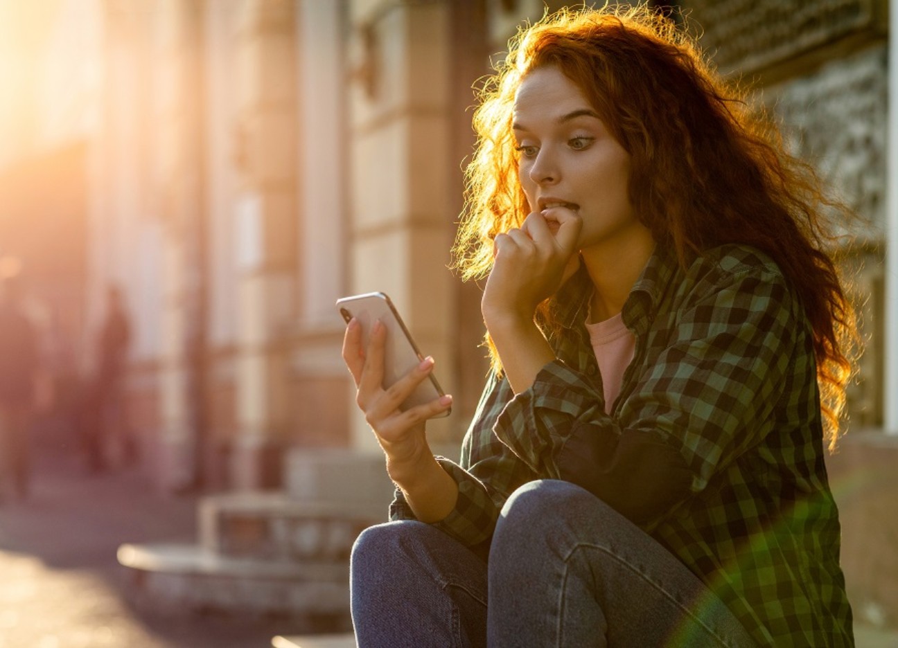 Bühnenbild Ratgeber Handy nervt? Eine junge Frau sitzt draußen auf einer Steinstufe und schaut gespannt in ihr Smartphone.