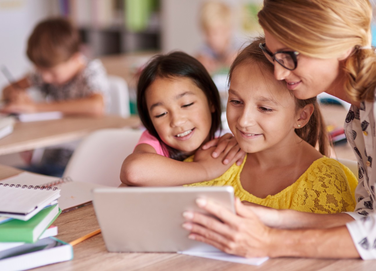 Eine Lehrerin zeigt zwei jungen Schülerinnen etwas auf dem Tablet. Sie sitzen in einem Klassenzimmer.