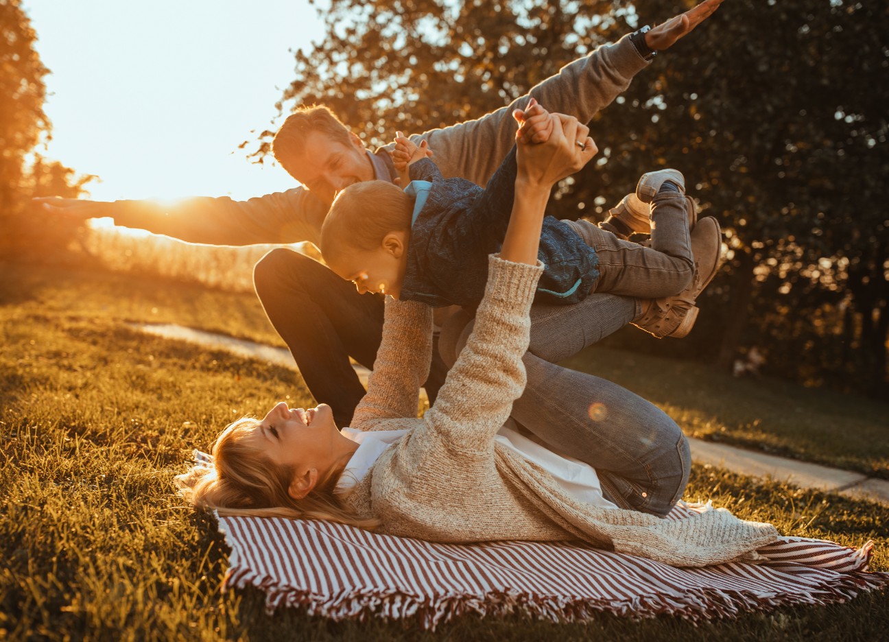 Bühnenbild Ratgeber Elterngeld Eltern albern zusammen mit ihrem jungen Kind auf einer Picknickdecke in der Sonne herum.