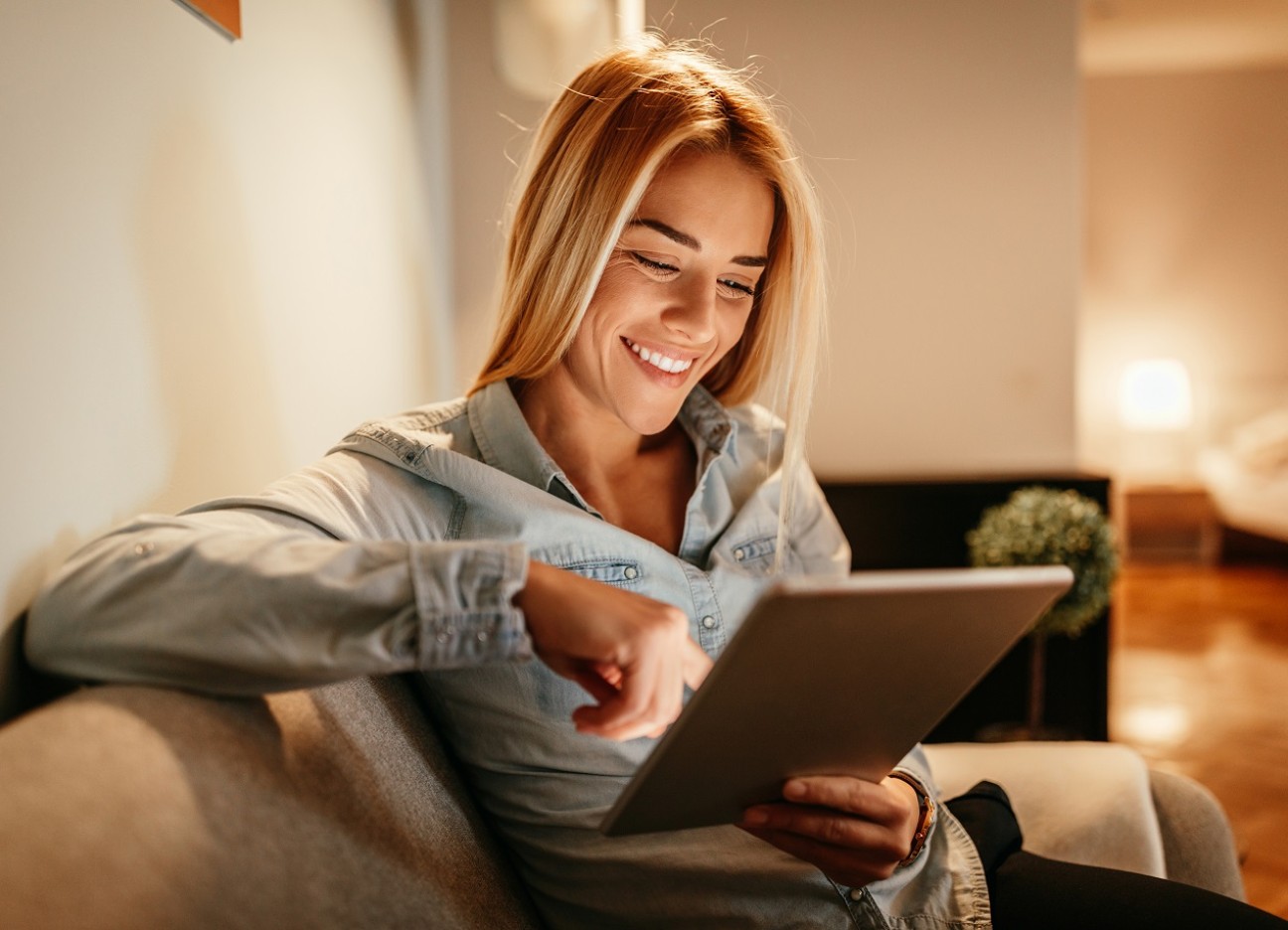 Bühnenbild Ratgeber Hausratversicherung Eine junge Frau lächelt, während sie etwas auf ihr Tablet durchliest. Sie sitzt in ihrem Wohnzimmer auf einer Couch.