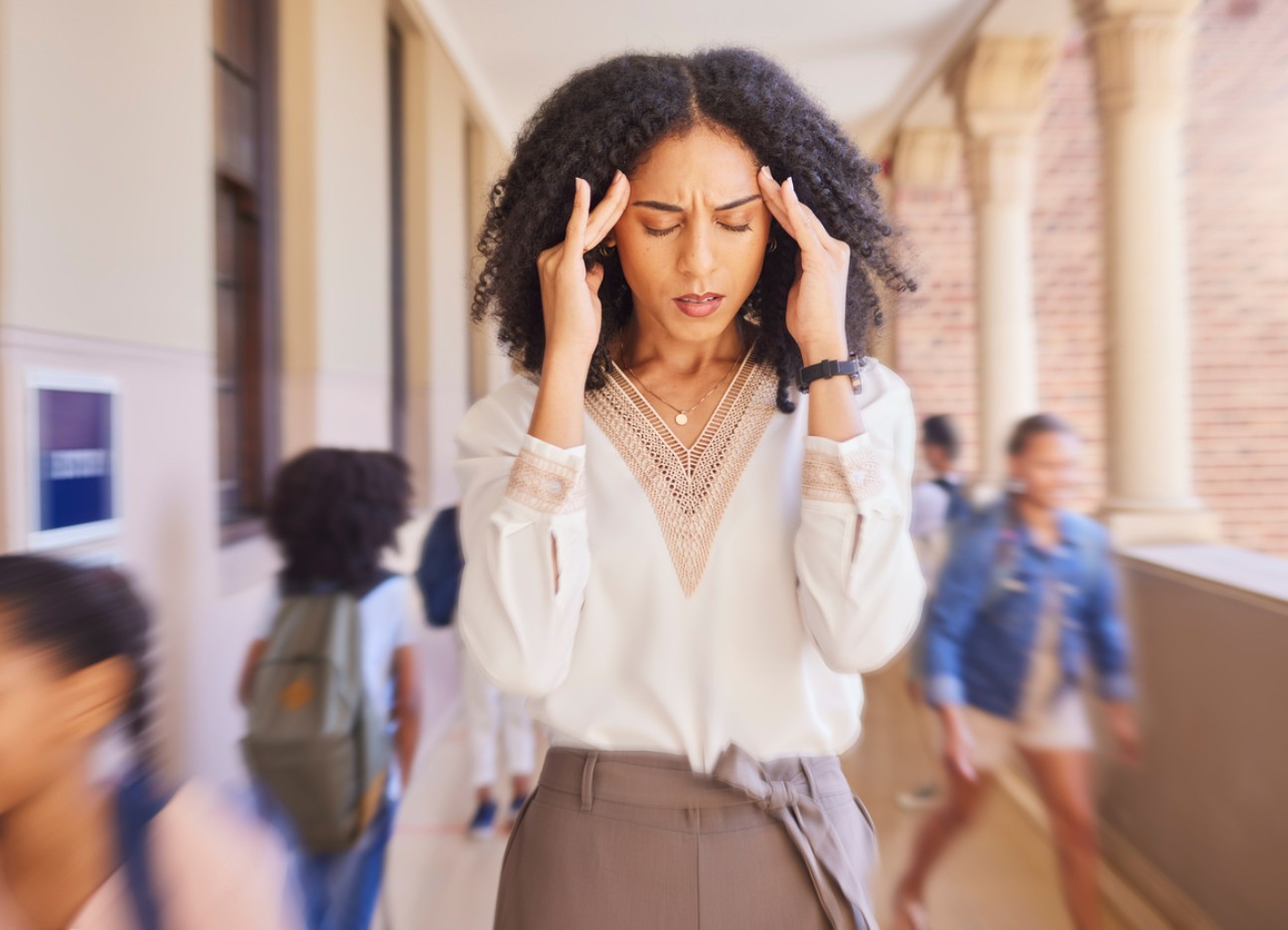 Bühnenbild Ratgeber Burnout Lehrer Eine Lehrerin steht im Schulflur und hält sich den Kopf. Es laufen mehrere Kinder um sie herum.