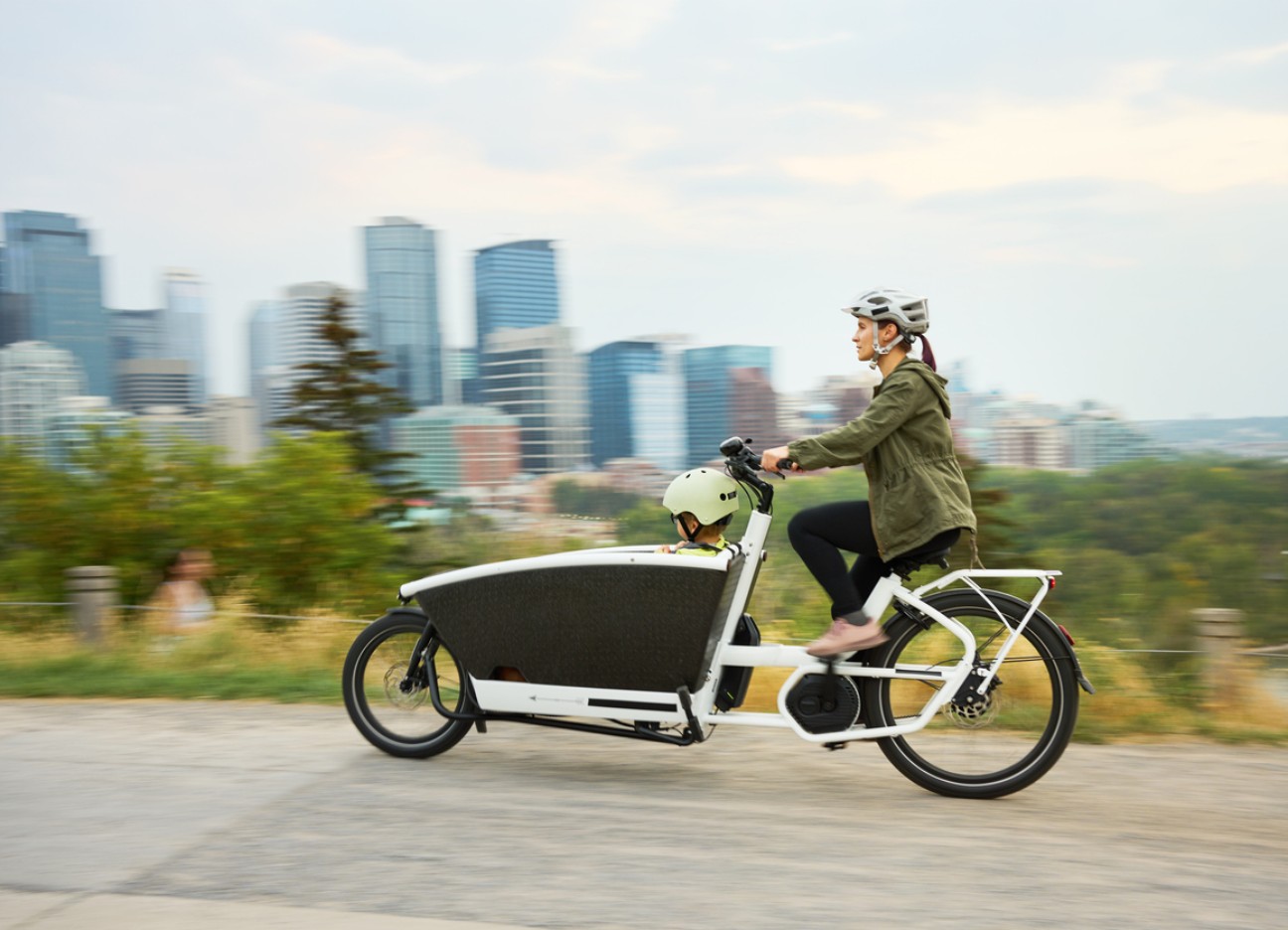 Bühnenbild Ratgeber Alternativen zum eigenen Auto Eine Mutter fährt ein Lastenrad, in dem ein Kind sitzt. Beide tragen einen Helm.