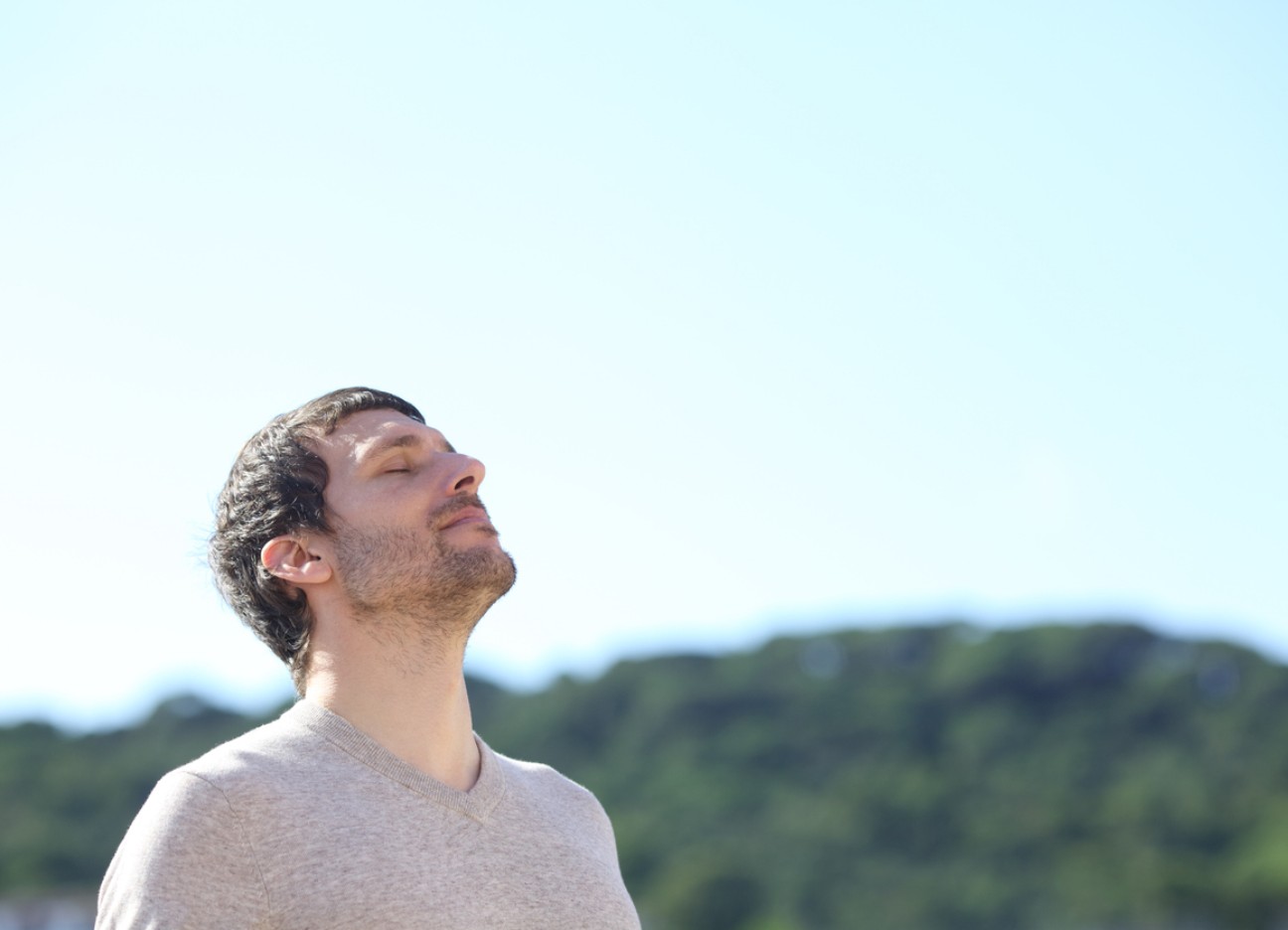 Ein Mann steht draußen mit geschlossenen Augen und hebt das Gesicht leicht in die Sonne, im Hintergrund eine grüne Hügellandschaft.