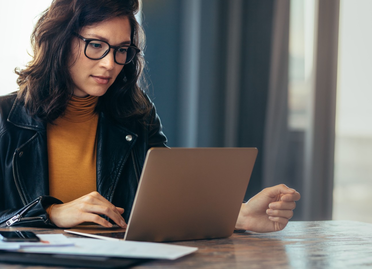 Eine Frau sitzt an einem Tisch und schaut auf ihren Laptop. Neben ihr liegen Dokumente. 