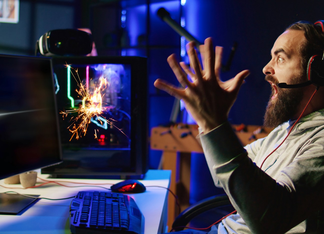 Ein Gamer sitzt am Schreibtisch und guckt erschrocken, weil aus seinem Computer Funken sprühen.