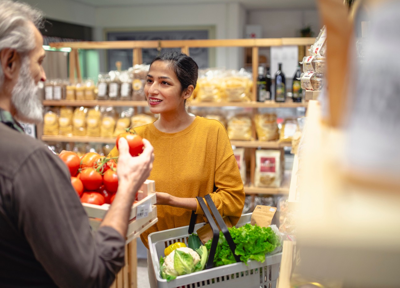 Eine junge Frau lächelt, während sie mit einem älteren Mann in einem Laden spricht und dabei einen Korb mit frischen Lebensmitteln hält. Im Hintergrund sind Regale mit verpackten Produkten zu sehen.