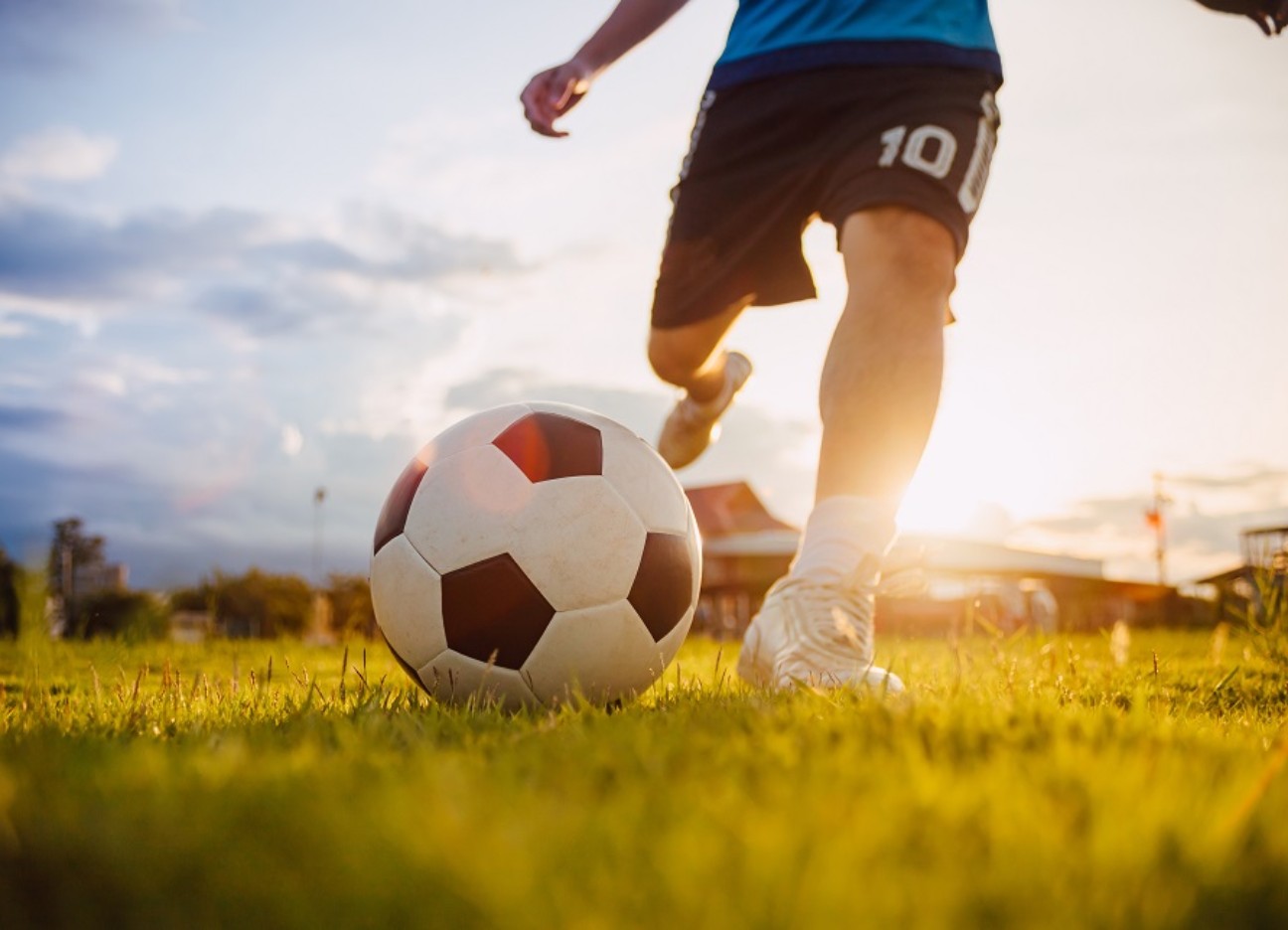 Ein Mann in Fußballklamotten (lediglich der Unterkörper sichtbar) schießt einen Fußball. Drum herum scheint die Sonne. 