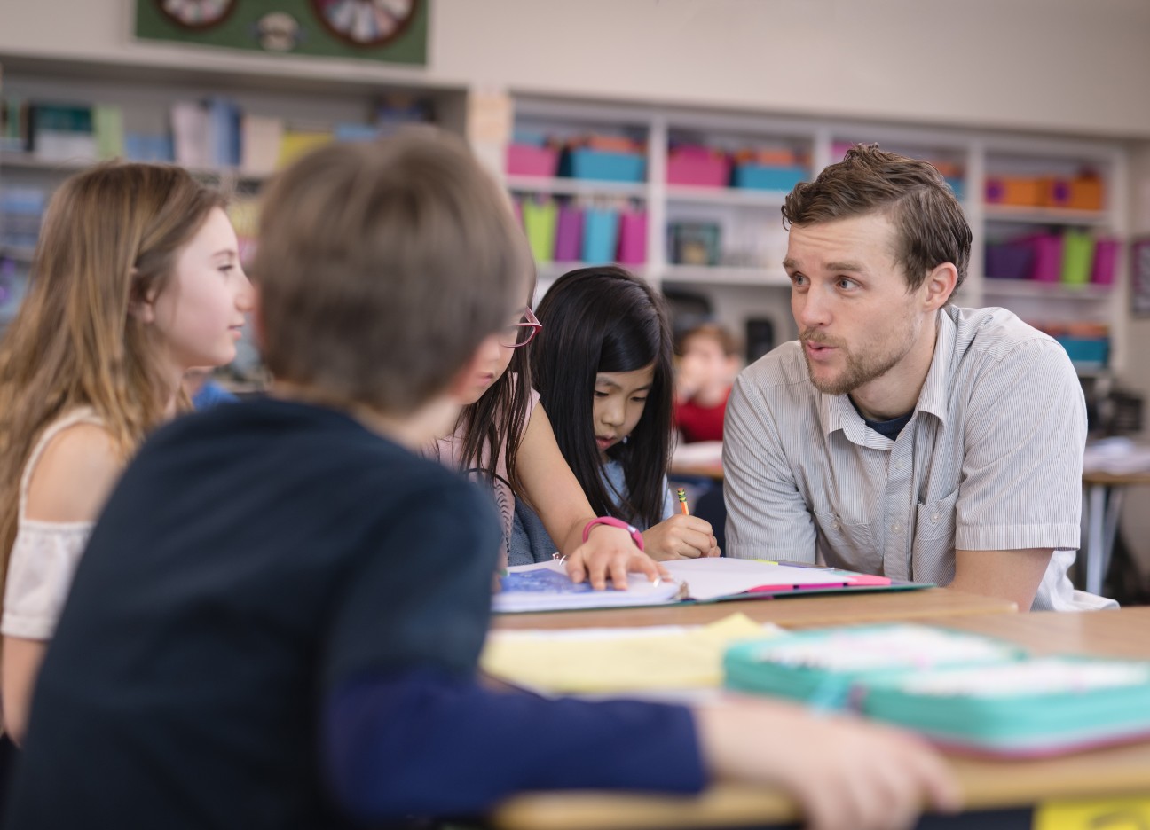 Bühnenbild Ratgeber Die verschiedenen Lehrerstadien Ein junger Lehrer unterhält sich mit zwei Schülern. Sie sitzen zu dritt an einem Tisch in einer Bibliothek.