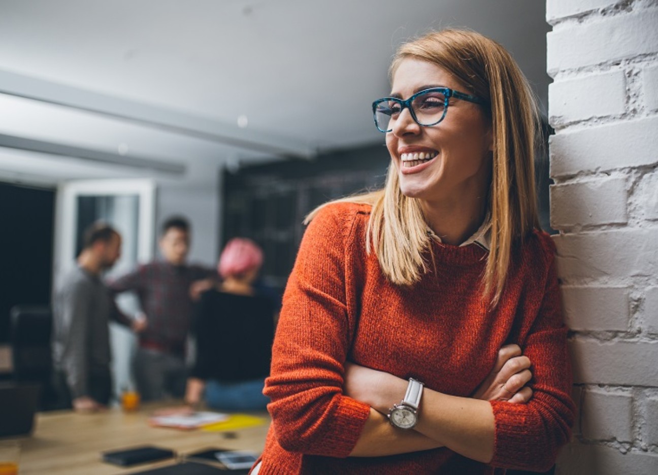 Female Investors - Frauen in den AR Person in rotem Pullover lehnt an einer weißen Wand, im Hintergrund sind weitere Personen in einem Büro.