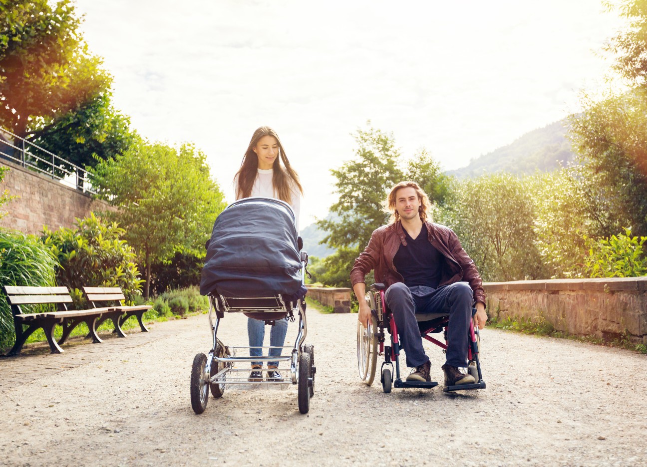 Grundfähigkeitsversicherung – wann ist sie nützlich? Eine Frau schiebt einen Kinderwagen, während ein Mann daneben in einem Rollstuhl sitzt. Sie spazieren gemeinsam einen Weg entlang.