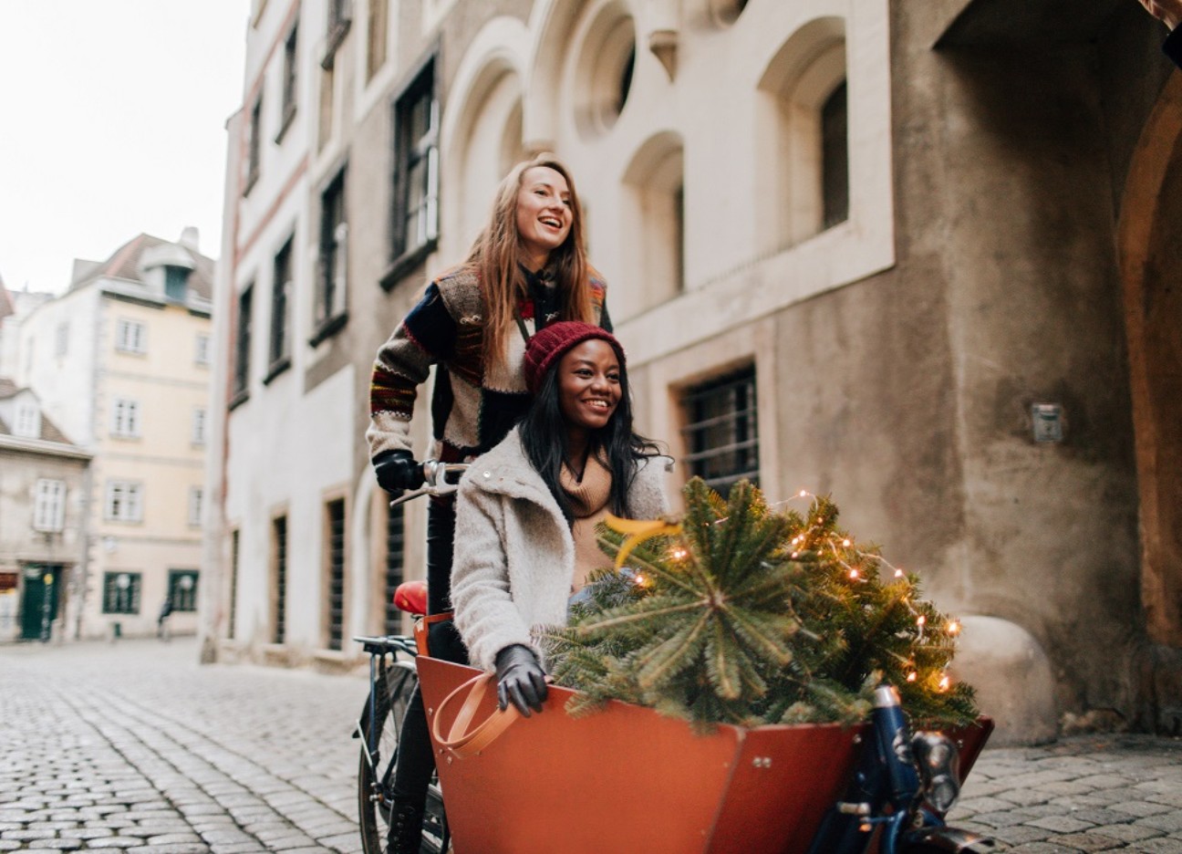 Bühnenbild Ratgeber Lastenrad E-Bikes winterfest Zwei Freundinnen sind zusammen mit einem Lastenrad durch die Straßen unterwegs. Eine Person fährt das Rad, die andere Person sitzt drin. Sie transportieren einen kleinen Nadelbaum.