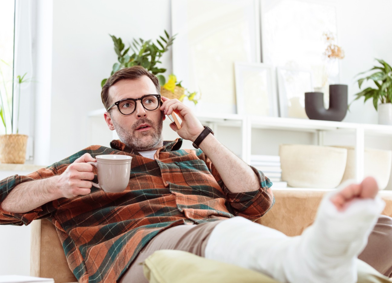 Ein Mann mit Brille sitzt auf einem Sofa, telefoniert und hält eine Tasse. Sein Bein ist in Gips und hochgelegt.